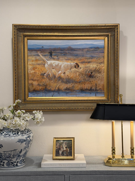 At Point framed sporting oil painting hung above a console styled with a brass lamp, blue-and-white jar, white flowers, and books