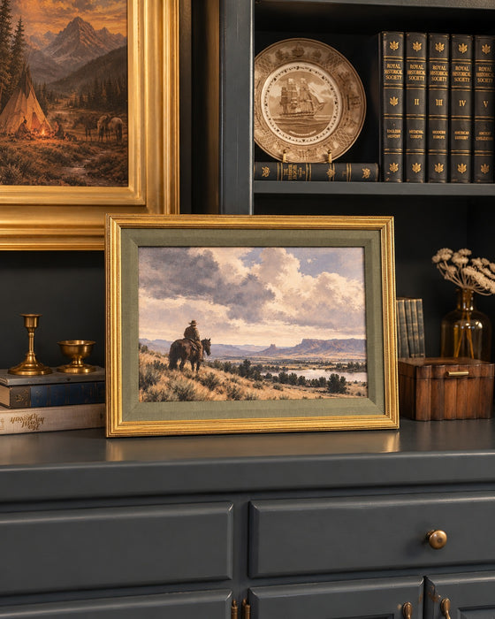 The Long View framed cowboy landscape with sage matting and gold frame, styled on a dark painted built-in beside brass candlesticks, books, and a larger framed painting